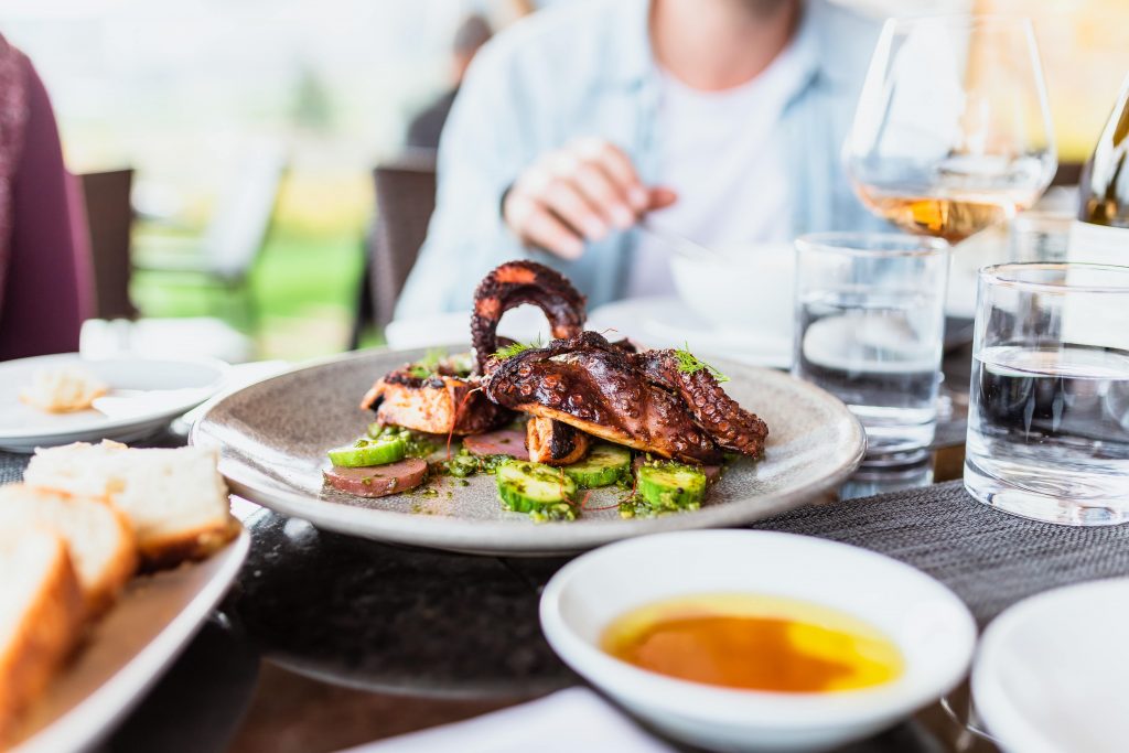 Close up of a plate on a restaurant table, surrounded by people. There is grilled octopus on the plate.